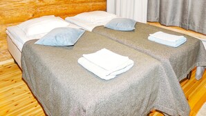 Une chambre avec deux lits, chacun recouvert d’une couverture grise et agrémenté d’oreillers blancs. On aperçoit un mur en bois et un rideau gris à l’arrière-plan.