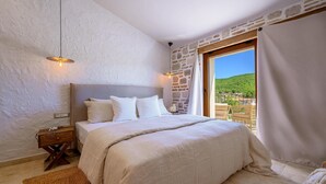 A bedroom with a stone wall, a bed with white bedding, a nightstand, a woven rug, and a view of greenery through a window. - Gorgeous home in Cera with WiFi (Cera)