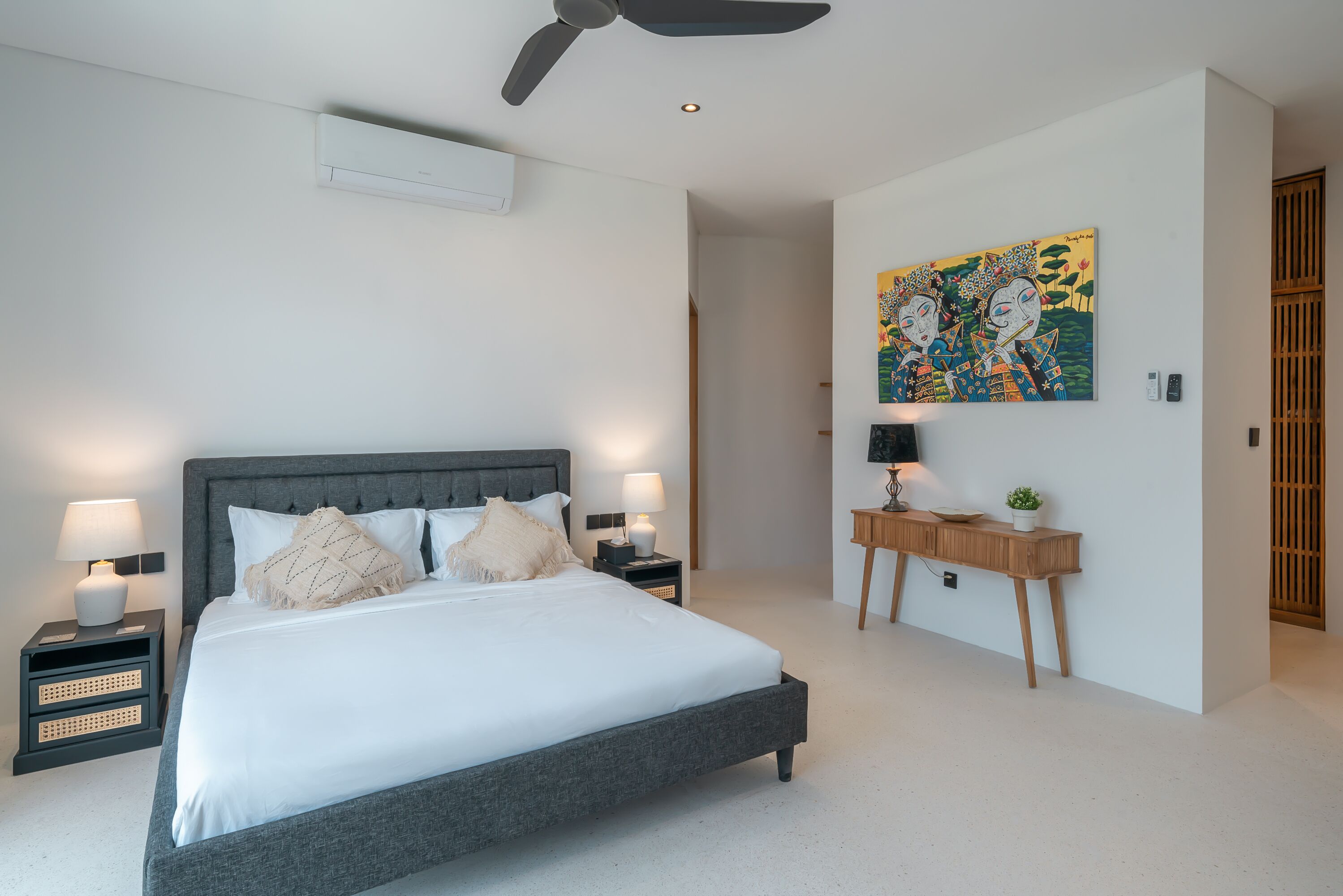 A modern bedroom with a large bed, bedside tables with lamps, a ceiling fan, a wall-mounted air conditioner, and a wooden nightstand with a plant.