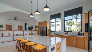 Eine moderne Küche mit einer großen Kochinsel, Holzbarhocker, Pendelleuchten und großen Fenstern mit Blick ins Freie.