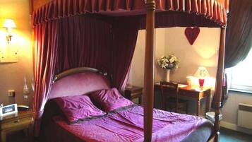 A four-poster canopy bed with a purple bedspread and pillows, a bedside table with a lamp, and a desk with a chair.