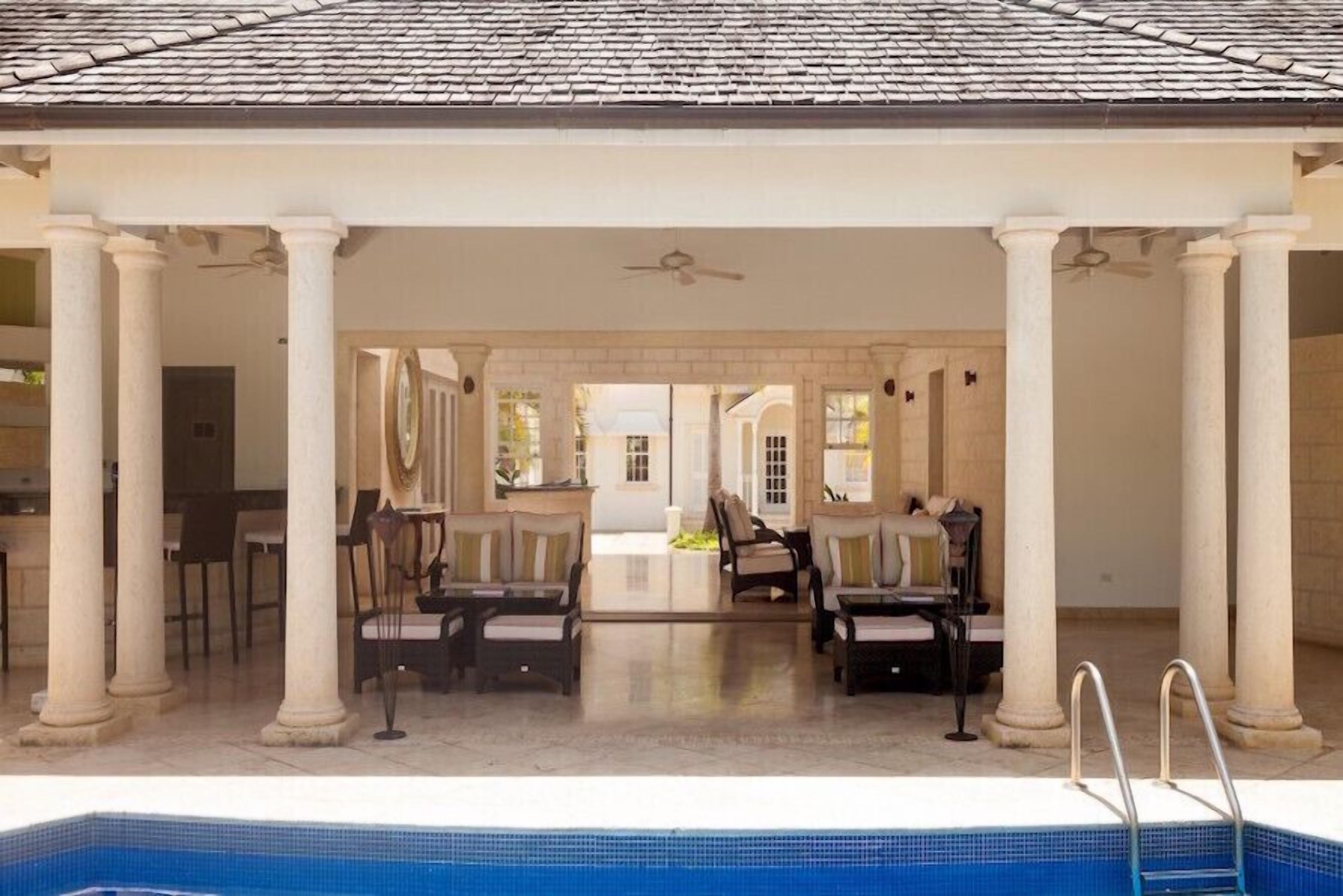 A covered outdoor seating area with a pool, columns, and a view of the interior.