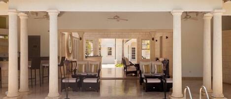 A covered outdoor seating area with a pool, columns, and a view of the interior.