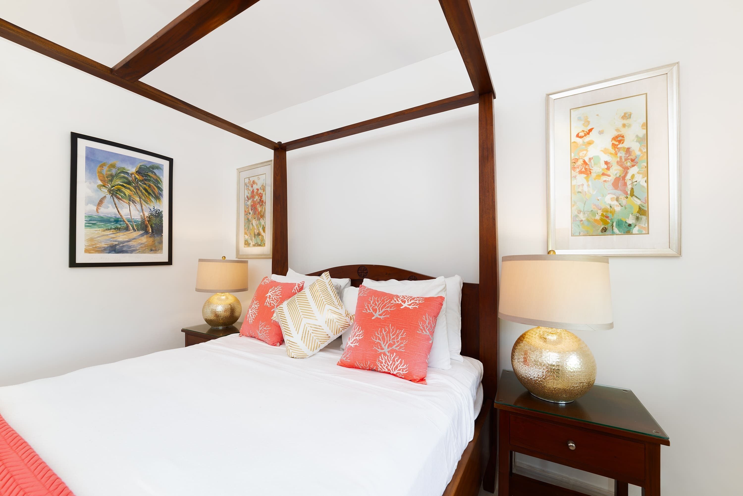 A four-poster bed with white linens and coral pillows, flanked by two bedside tables with lamps, in a room with wooden beams and framed artwork on the walls.