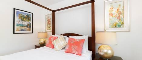 A four-poster bed with white linens and coral pillows, flanked by two bedside tables with lamps, in a room with wooden beams and framed artwork on the walls.