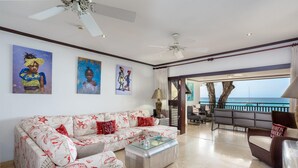 Un salon avec un canapé, une table basse et des œuvres d’art accrochées au mur. Il y a un balcon avec vue sur la mer.