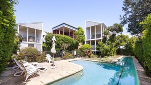 A swimming pool with lounge chairs and umbrellas, surrounded by greenery and modern buildings.