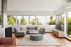 A modern living room with a sectional sofa, a round ottoman, and a coffee table. There is a TV on a wooden stand and a large window with a view of greenery.