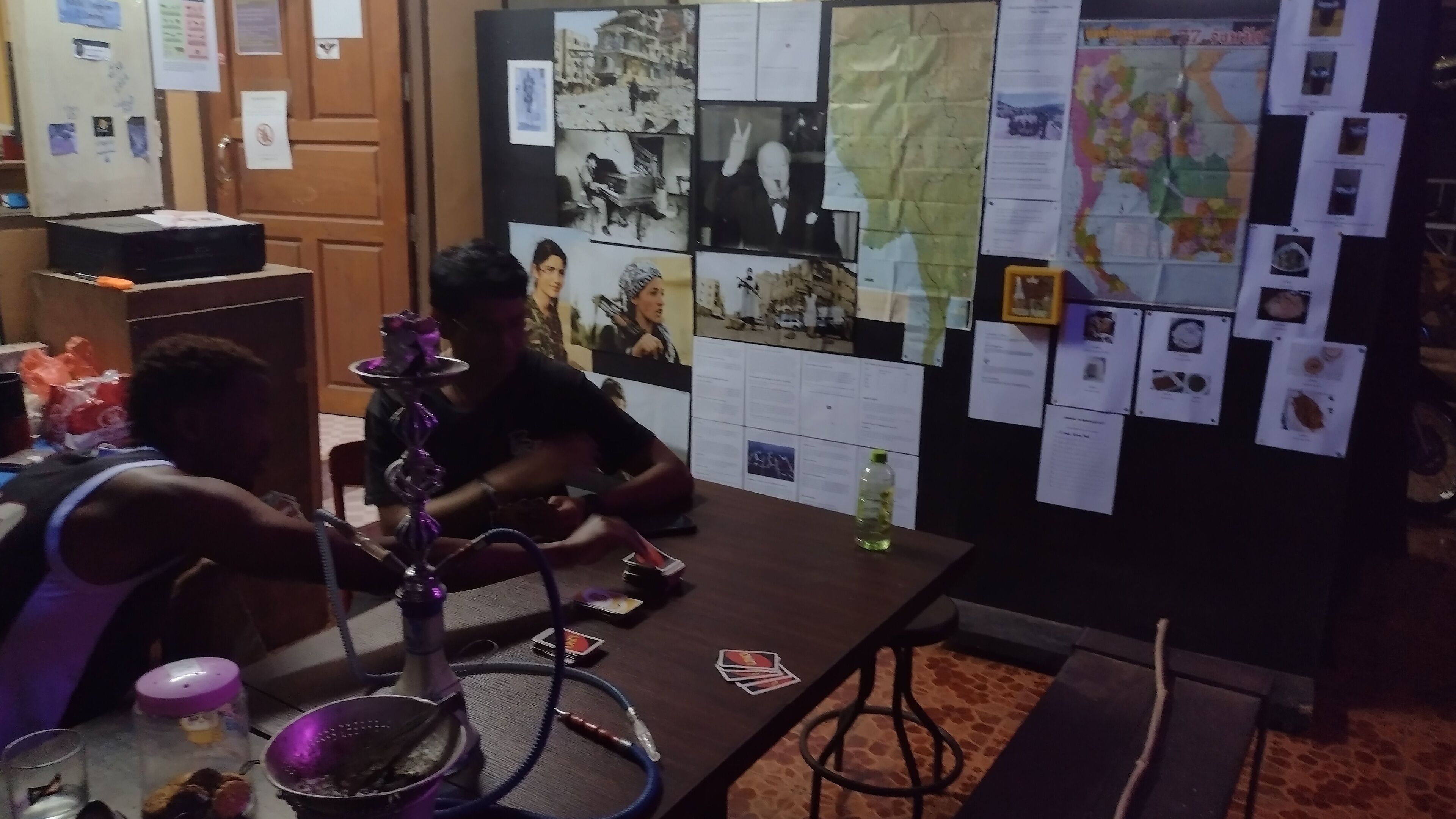 Una habitación con un escritorio de madera, un narguilé y un tablón de anuncios con varios papeles y fotografías.