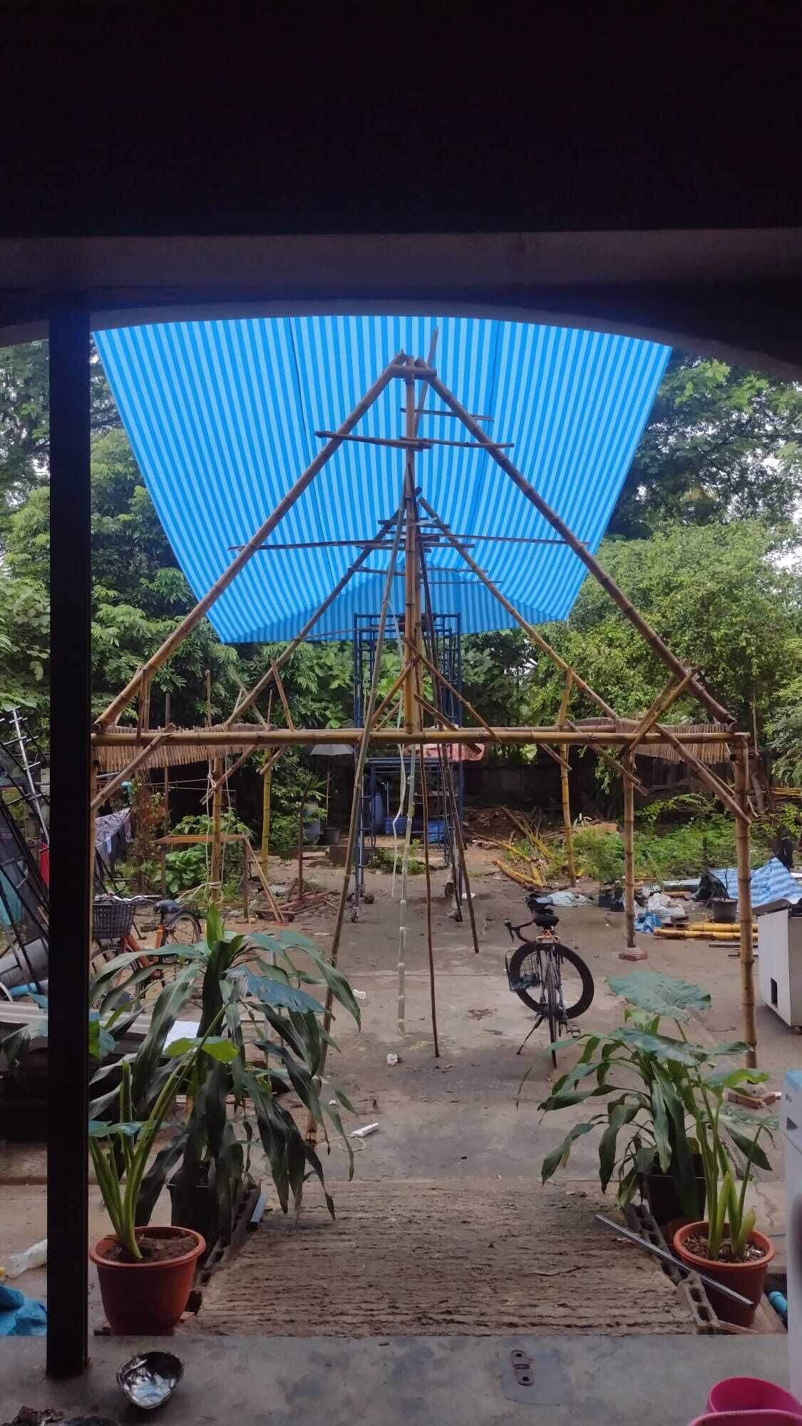 A bamboo structure with a blue tarp roof, surrounded by potted plants and bicycles.