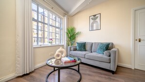 Un salon avec un canapé gris, une table basse ronde en verre sur laquelle sont posés des livres et un vase, une plante en pot et une fenêtre avec des rideaux.