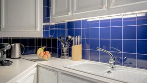 Une cuisine avec des meubles blancs, un plan de travail recouvert de carreaux bleus, une bouilloire en acier inoxydable, une machine à café, un panier à fruits et un bloc à couteaux.