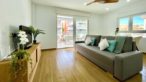 A modern living room with a grey sofa, wooden flooring, a ceiling fan, and a view of the outside balcony.