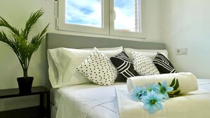A neatly made bed with white bedding and decorative pillows, a potted plant, and a window with a view of the sky.