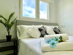 Ein ordentlich bezogenes Bett mit weißer Bettwäsche und dekorativen Kissen, eine Topfpflanze und ein Fenster mit Blick zum Himmel.