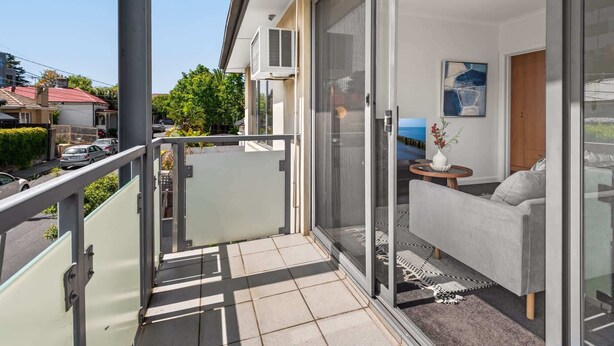 Un balcon avec une porte-fenêtre coulissante, un canapé gris, une petite table ronde avec un vase, et une vue sur la rue et les maisons.