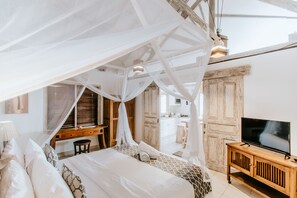 A bedroom with a canopy bed, a wooden desk, a television, and a bathroom visible through an open door.