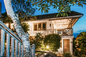A two-story house with a balcony, surrounded by lush greenery and a view of the ocean.