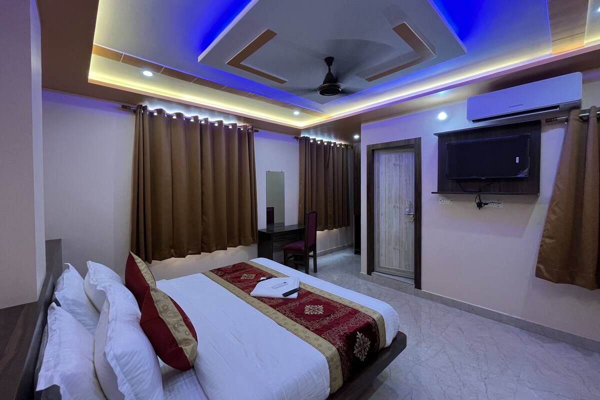 A hotel room with a bed, curtains, a desk, a chair, a television, and an air conditioning unit.