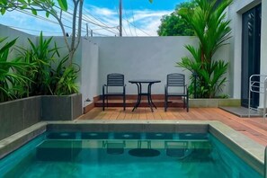 Una piscina con dos sillas y una mesa, rodeada de plantas y una pared blanca.