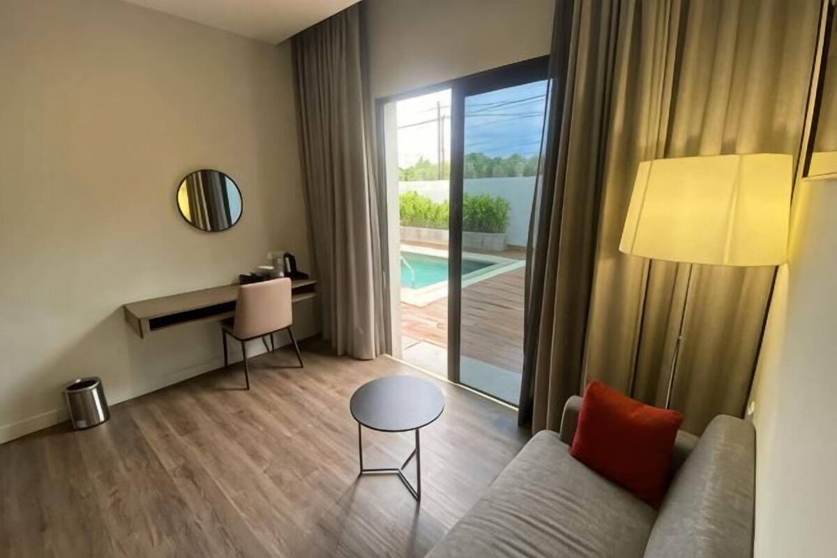 A hotel room with a sofa, a small table, a desk with a chair, a lamp, and a view of a swimming pool through a glass door.