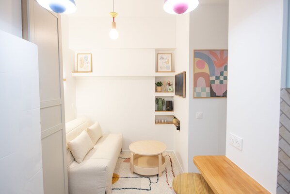 A modern, minimalist living room with a white sofa, a small round table, and a wooden bench. There are shelves with decorative items and framed pictures on the wall. - Charmant Studio Pour Deux Près de L'arc de Triomphe (Paris)