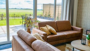 A modern living room with a leather sofa, a round coffee table, and a view of a green field through large windows. - Villa in Leons near Frisian Lakes (Leons)