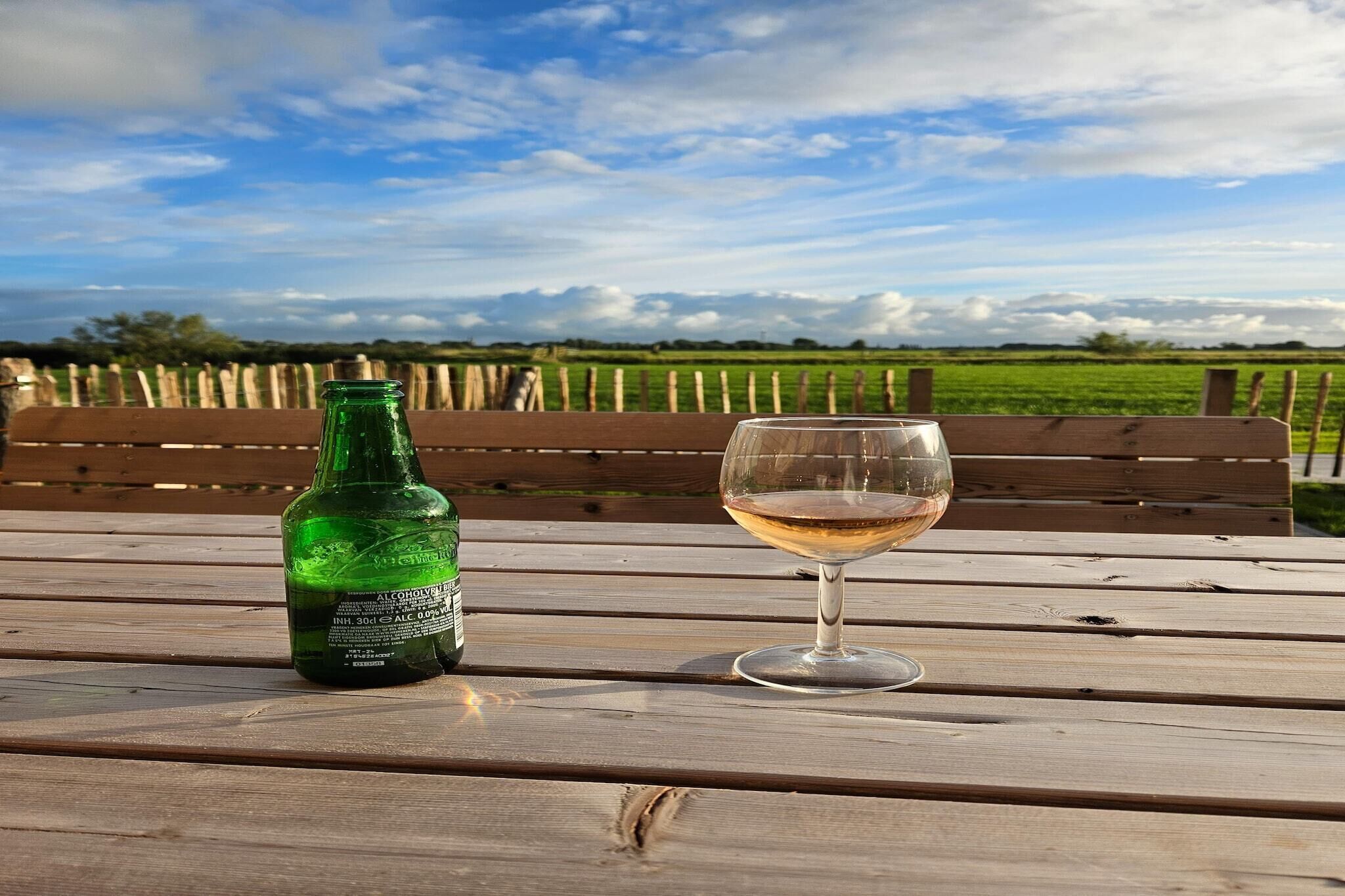 A bottle of beer and a glass on a wooden table with a bench and a view of a field and sky.