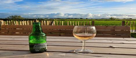 A bottle of beer and a glass on a wooden table with a bench and a view of a field and sky.