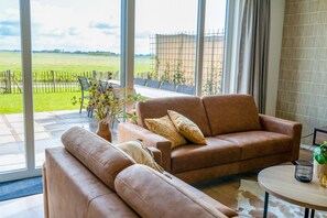 Een moderne woonkamer met een leren bank, een ronde salontafel en uitzicht op een groen veld door grote ramen.