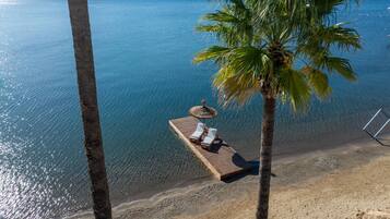 A wooden deck with two sun loungers and a straw umbrella, overlooking a clear blue sea with palm trees.