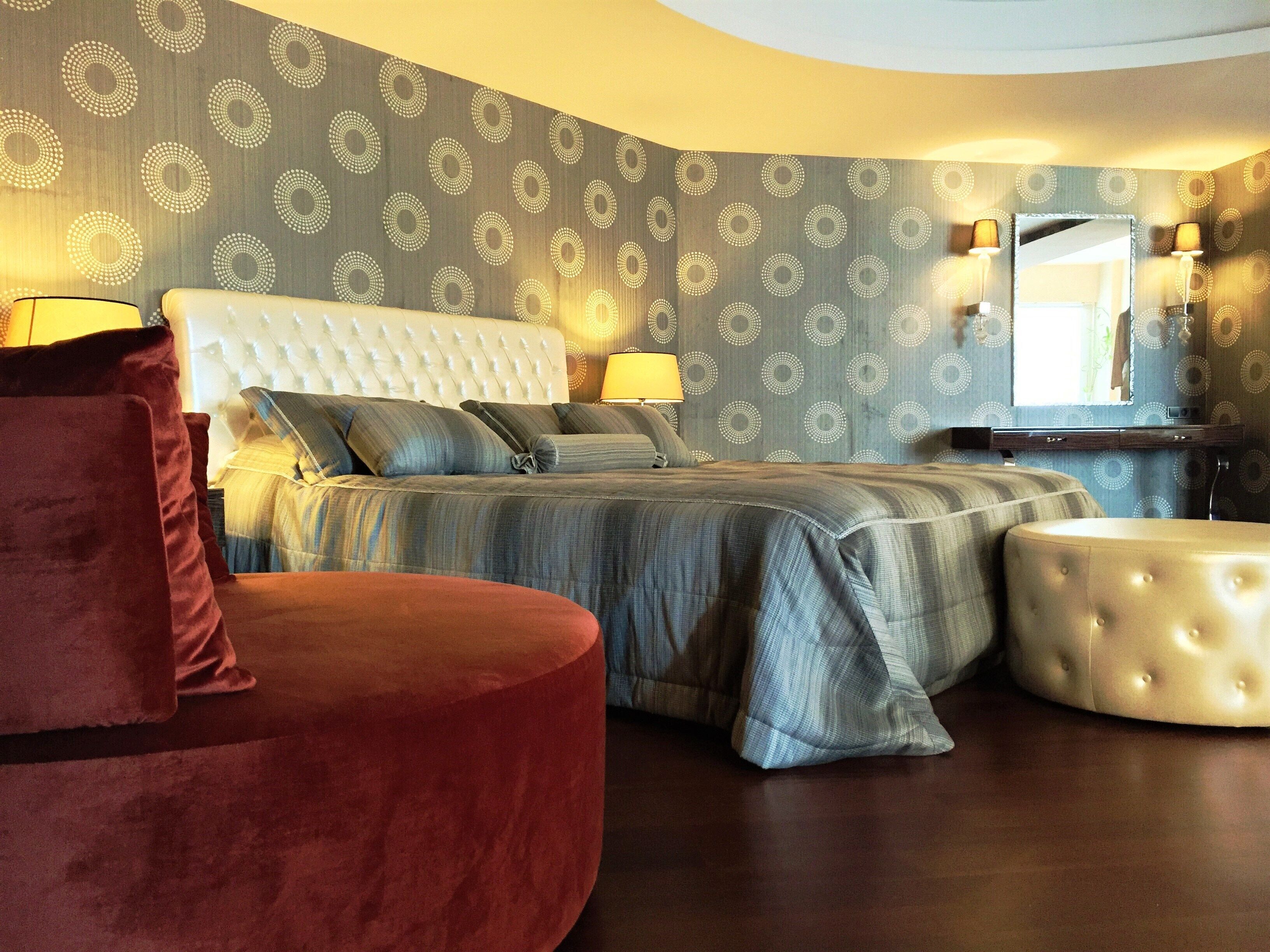 A bedroom with a tufted headboard, a bed with a striped bedspread, two bedside lamps, a round ottoman, and a red chair.