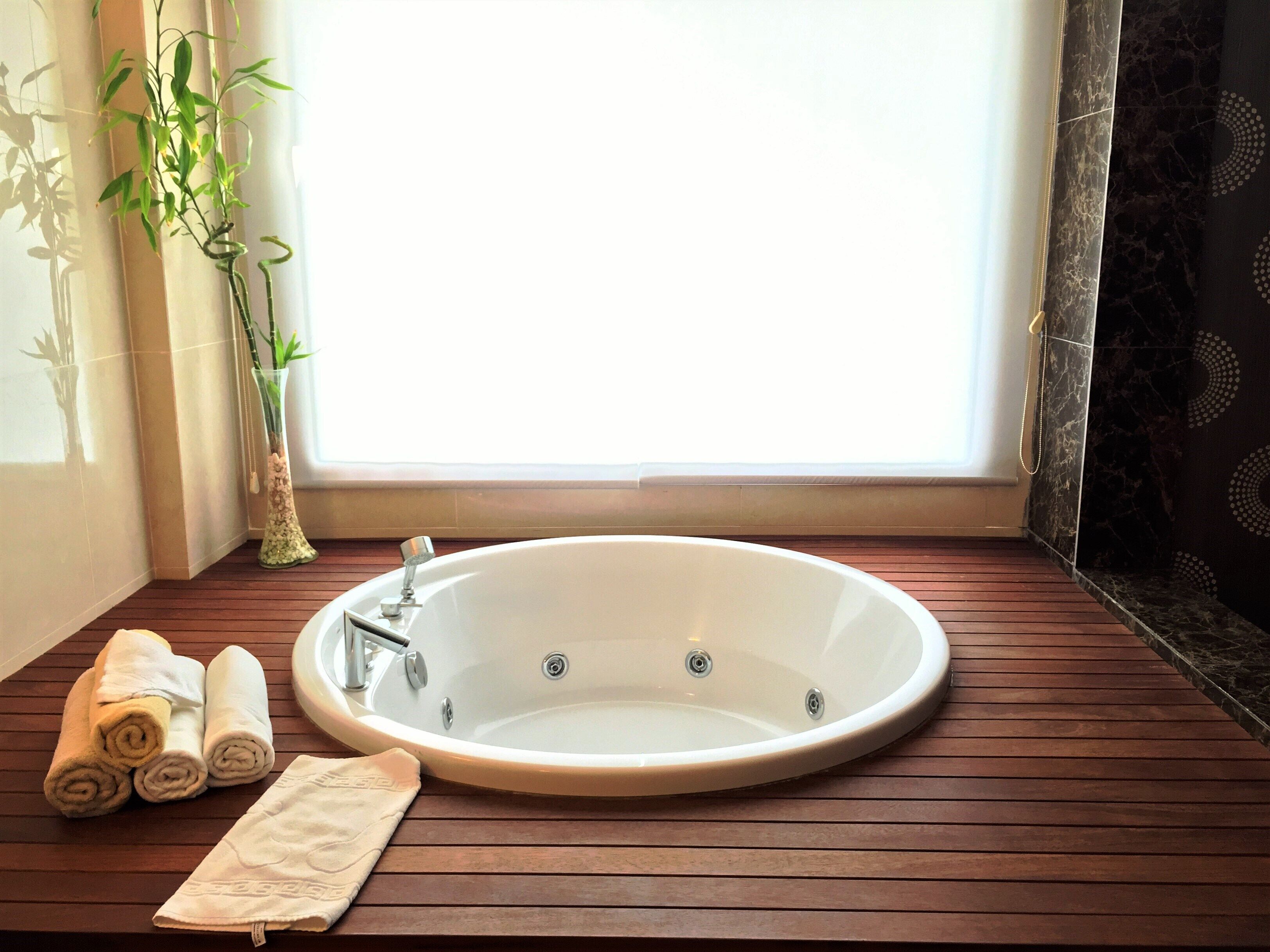 A bathroom with a large oval bathtub, wooden flooring, a window with curtains, and a potted plant.