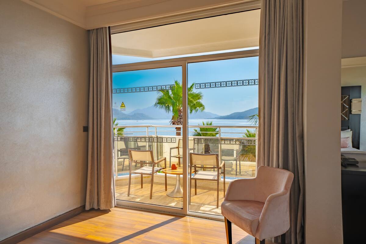 Una habitación con un gran ventanal corredero que da a un balcón con vistas al mar y a las montañas. Hay dos sillas y una mesita en el balcón.