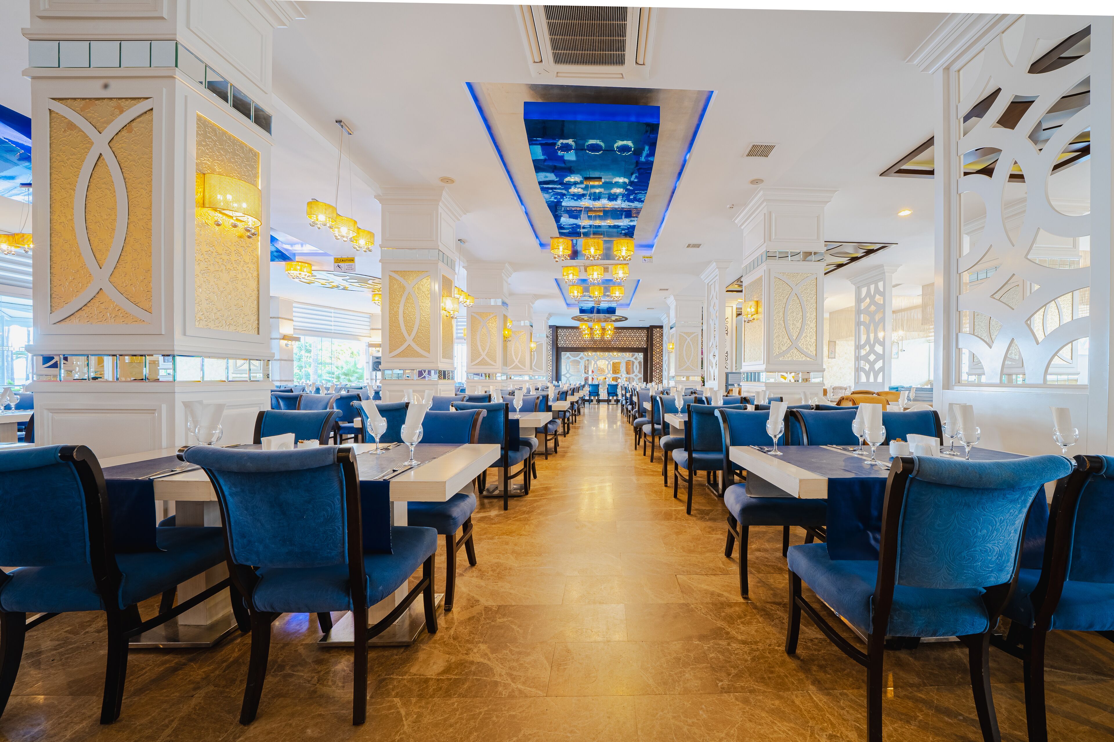 Un restaurant spacieux avec des chaises à revêtement bleu et des tables blanches, agrémenté d’un lustre central et de panneaux décoratifs au plafond.