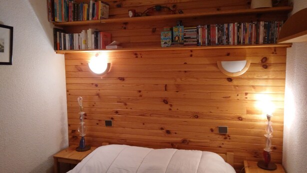 A wooden-paneled room with a bed, two bedside tables, and a bookshelf with books and decorative items.