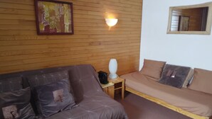 A room with a wooden wall, a bed, a sofa, a nightstand, and a wall-mounted lamp.