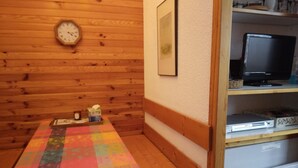 A room with a wooden wall, a wooden bench, a table with a colorful tablecloth, a clock, a framed picture, a television, and a white cabinet.