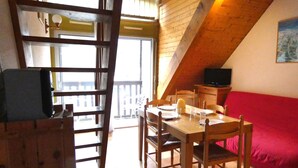 Un coin repas avec une table et des chaises en bois, un canapé rouge, un téléviseur et un balcon doté d’une balustrade.