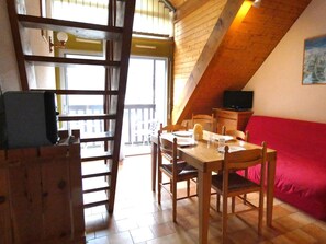 A dining area with a wooden table and chairs, a red sofa, a television, and a balcony with a railing.