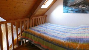 Une chambre avec un plafond en pente, un lit superposé en bois, une couverture colorée et un tableau représentant un paysage enneigé accroché au mur.
