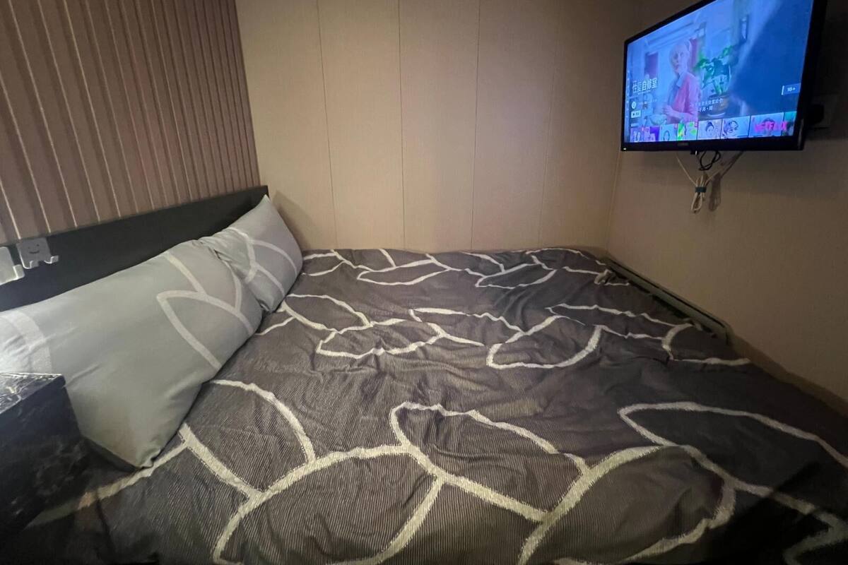 Une chambre d’hôtel avec un lit recouvert d’une couvre-lit à motifs, un téléviseur fixé au mur diffusant une émission et une fenêtre avec des stores.