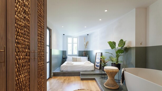 A modern bedroom with a large bed, a small bedside table, a potted plant, and a bathtub.