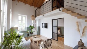 A spacious room with a high ceiling, wooden flooring, a staircase, a dining area with a table and chairs, and a bed with a view.