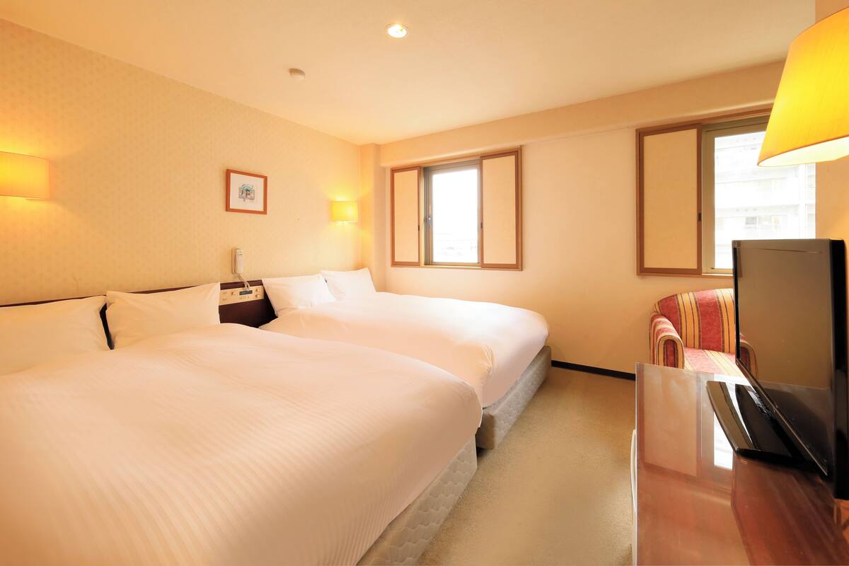 A hotel room with two beds, a desk, a chair, a TV, a window, and a lamp.