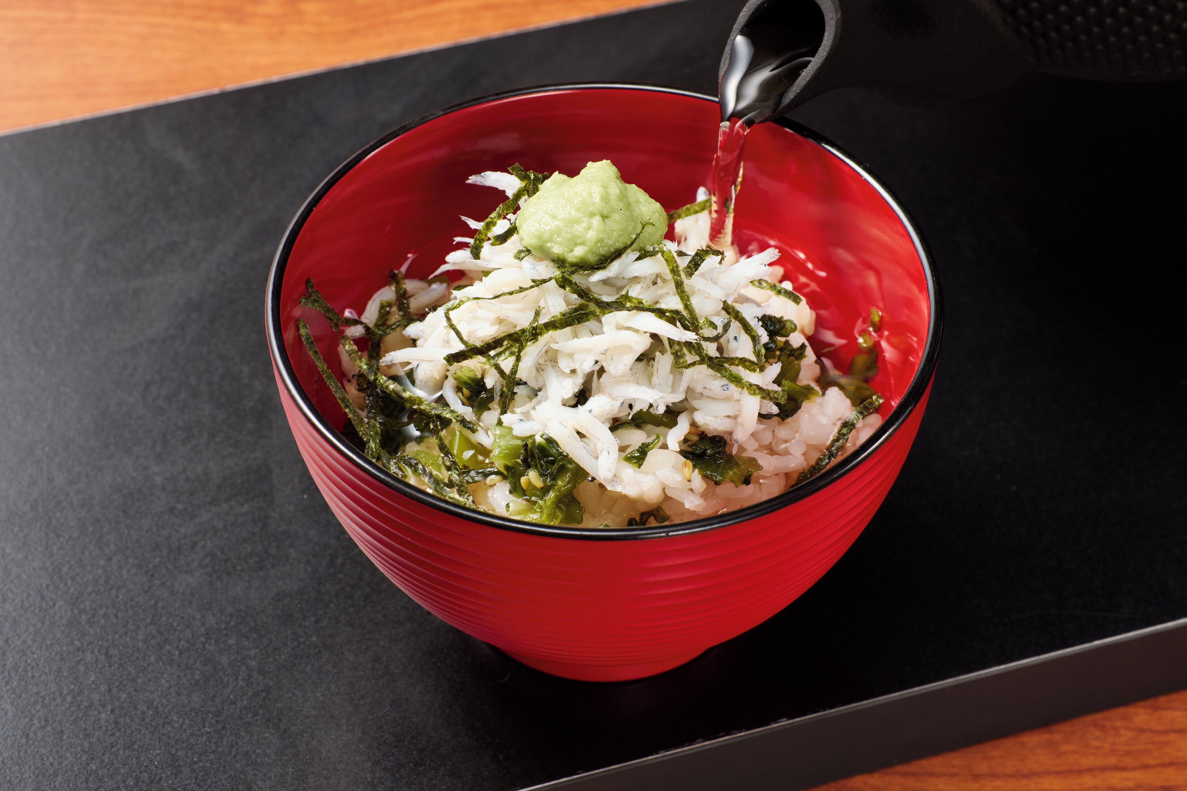 A red bowl with white rice, green vegetables, and a green sauce being poured into it.