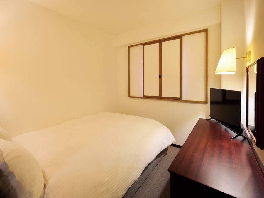 A hotel room with a bed, a desk with a flat-screen TV, a telephone, and wall-mounted lamps.