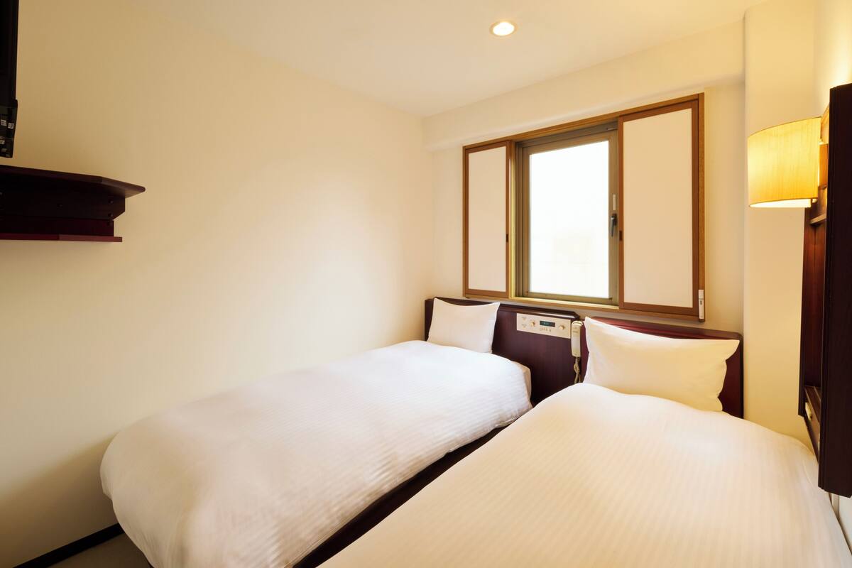 A hotel room with two single beds, a window with a view, a bedside table, and a wall-mounted lamp.
