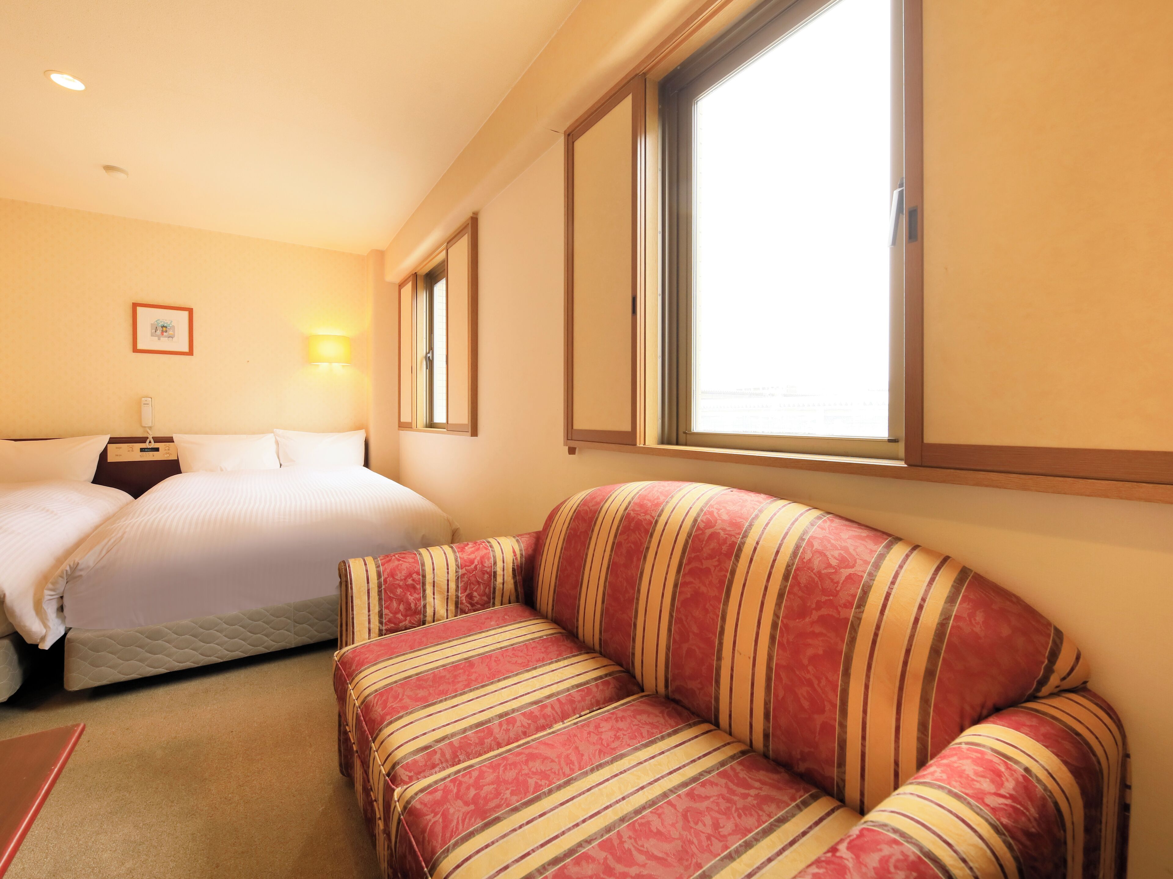 Een hotelkamer met twee bedden, een gestreept bankje, een nachtkastje met een lamp en een raam met uitzicht.