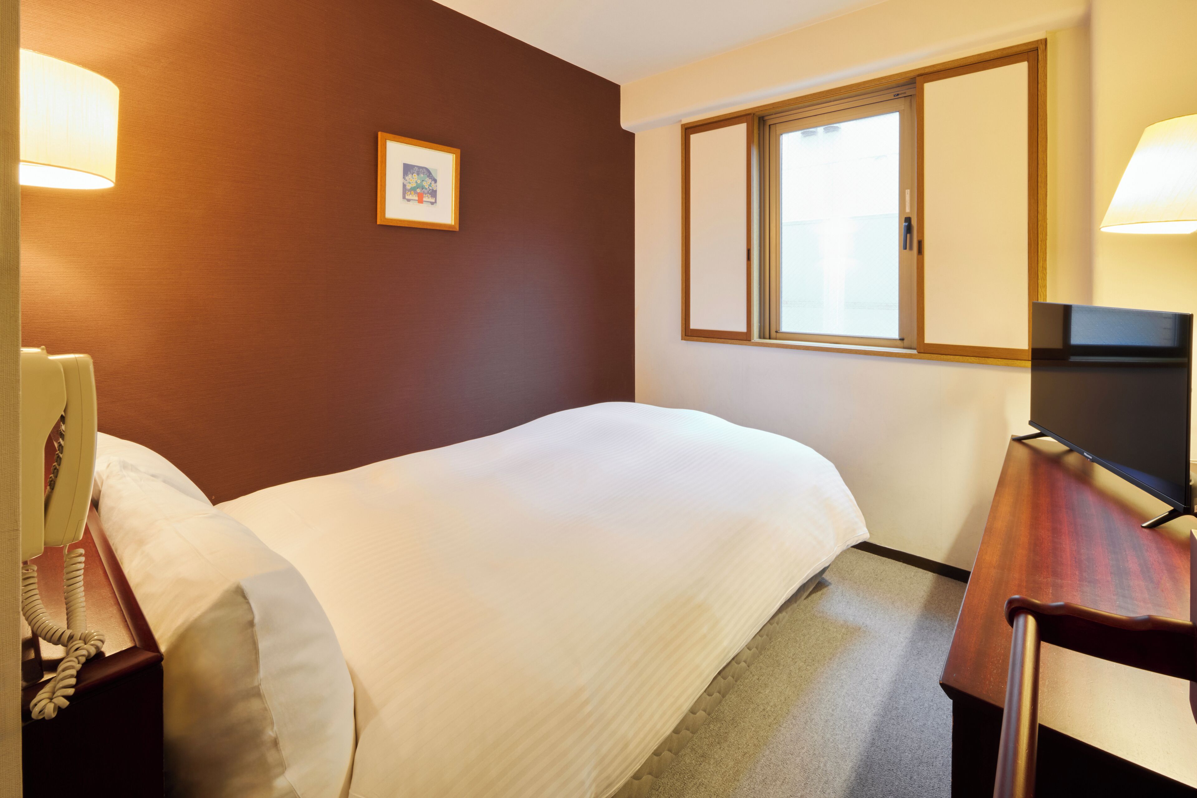 A hotel room with a bed, a desk with a lamp, a telephone, and a window with blinds.